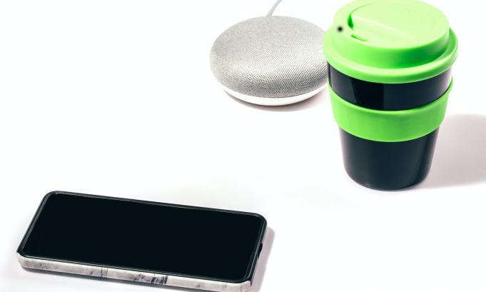 Smart speaker with smartphone and reusable coffee cup with green lid on white background.