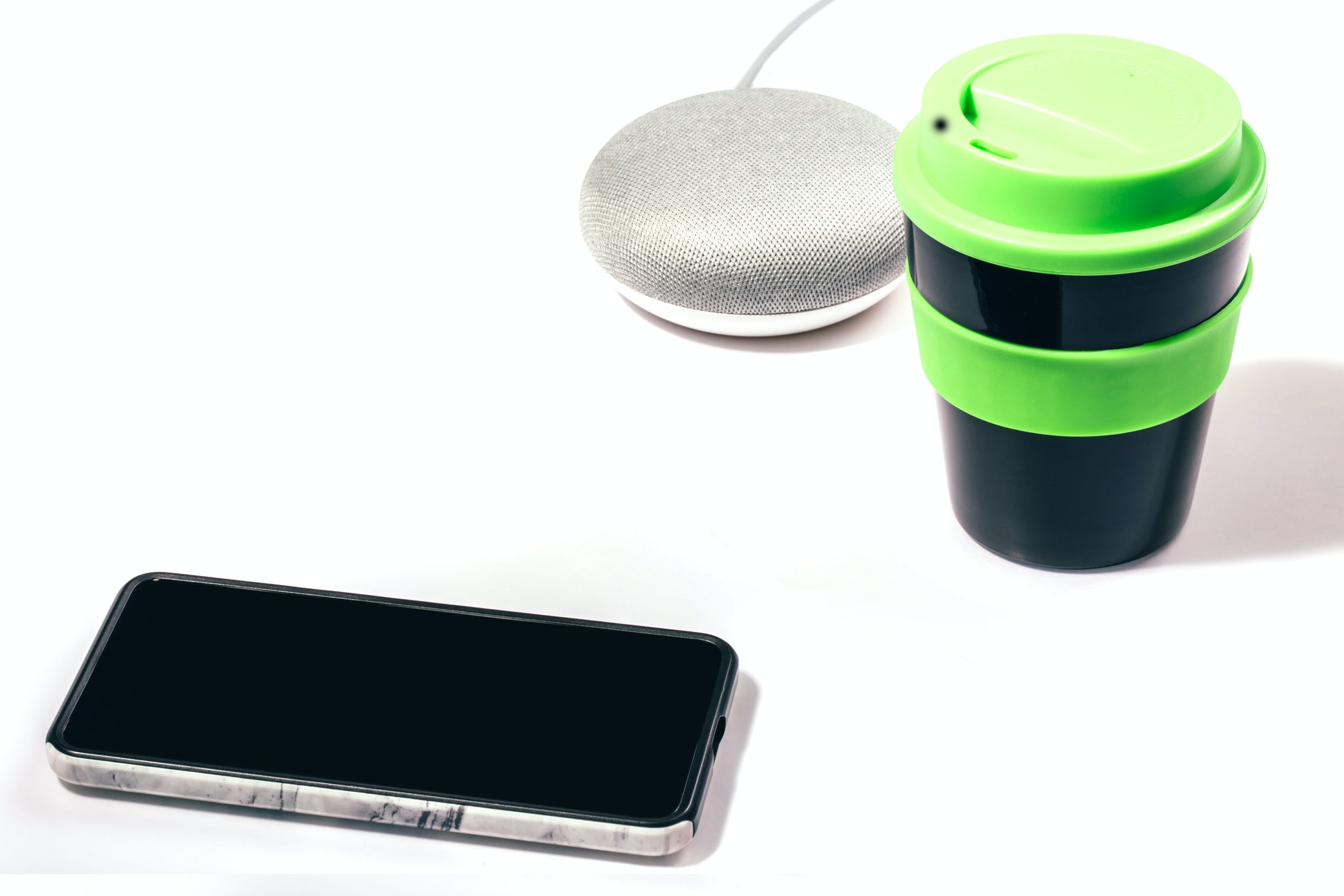 Smart speaker with smartphone and reusable coffee cup with green lid on white background.